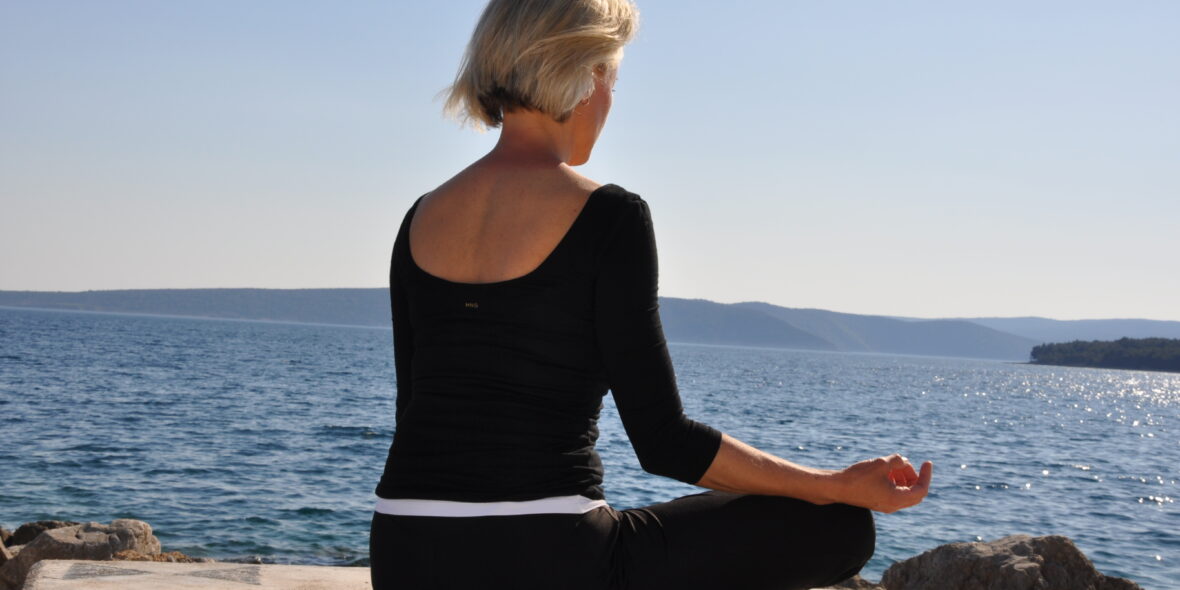 Sabine Barta in Meditationshaltung am Meer sitzend, blauer Himmel, Achtsamkeit in Therapie & Beratung