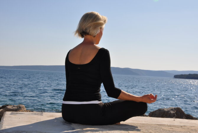 Sabine Barta in Meditationshaltung am Meer sitzend, blauer Himmel, Achtsamkeit in Therapie & Beratung